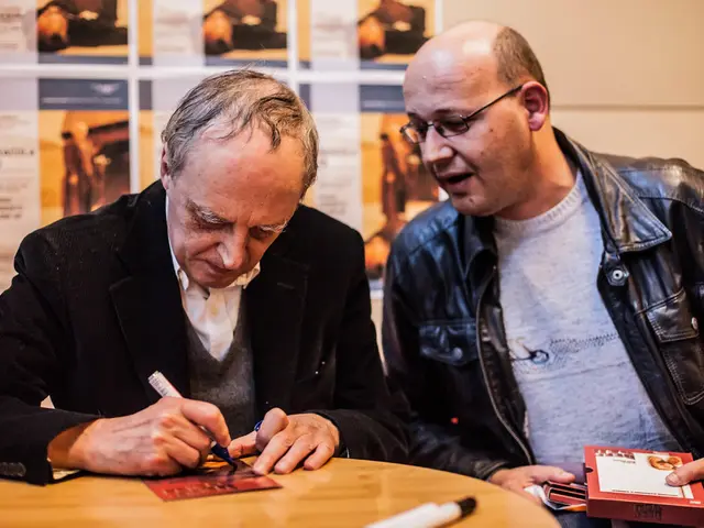 In this image there are two men who are sitting in the chairs. The man on the left side is writing...